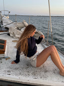 A woman with long light brown hair sits on the deck of a sailboat, wearing a dark navy long-sleeve top and white striped shorts, with the ocean and sky in the background.